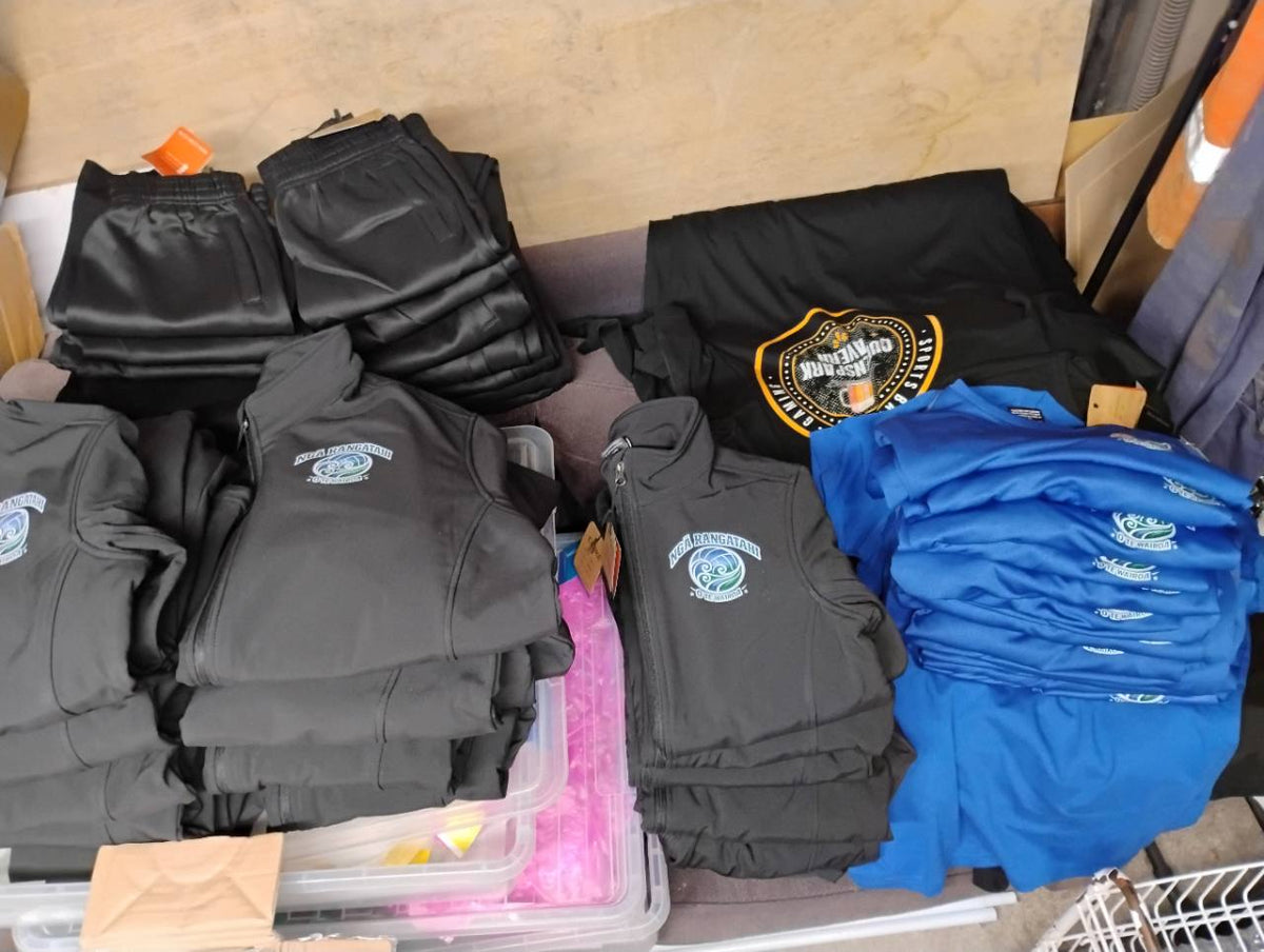 Stacks of grey zip jackets and blue T-shirts with matching white chest logos, alongside folded black garments.