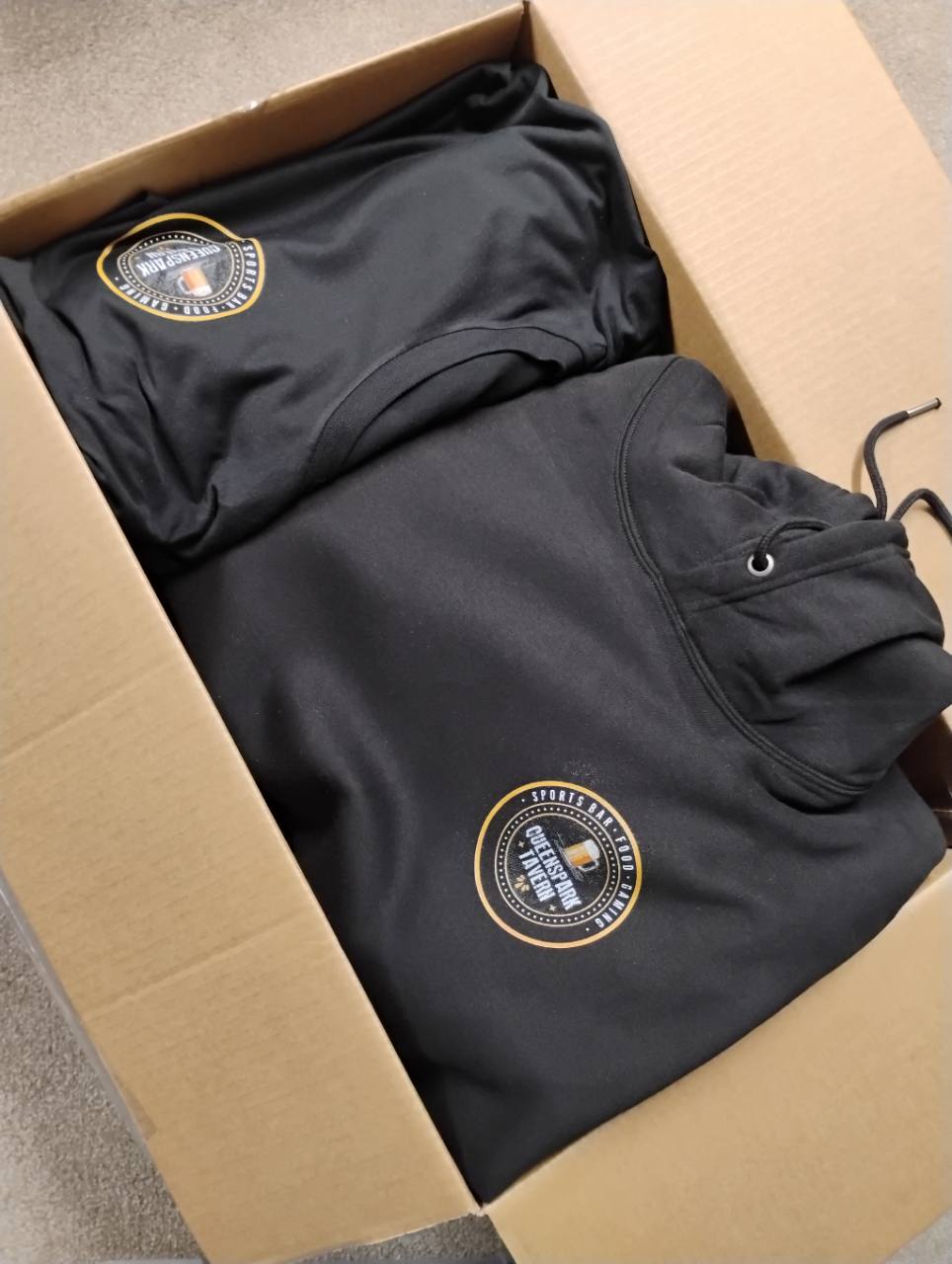 Black hoodie and T-shirt packed in a box, each with a small white-and-yellow Queenspark Tavern chest logo.