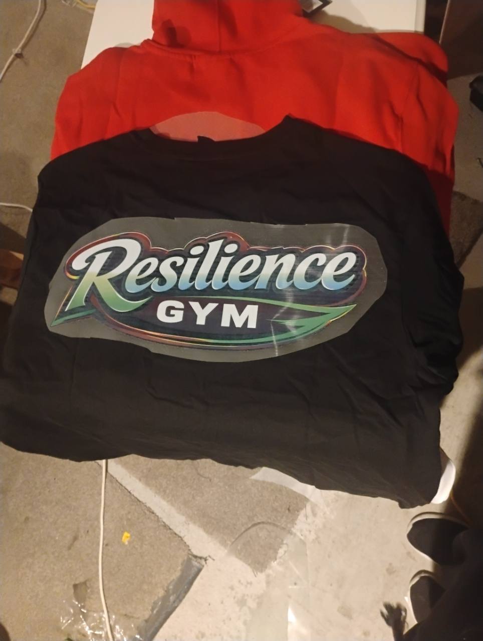 Black garment with a multicolour Resilience Gym logo transfer laid across it, with a red hoodie behind.
